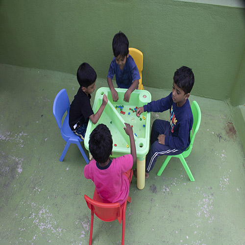 four children are playing with the beads in the table and are sitting in the chairs.